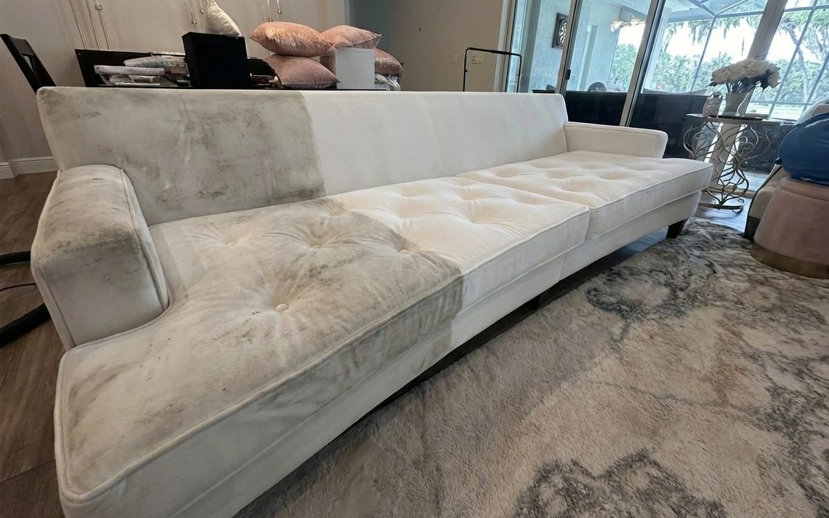 A white sofa photographed mid-job: the left half is gray with soil, the right half is bright and clean after steam extraction